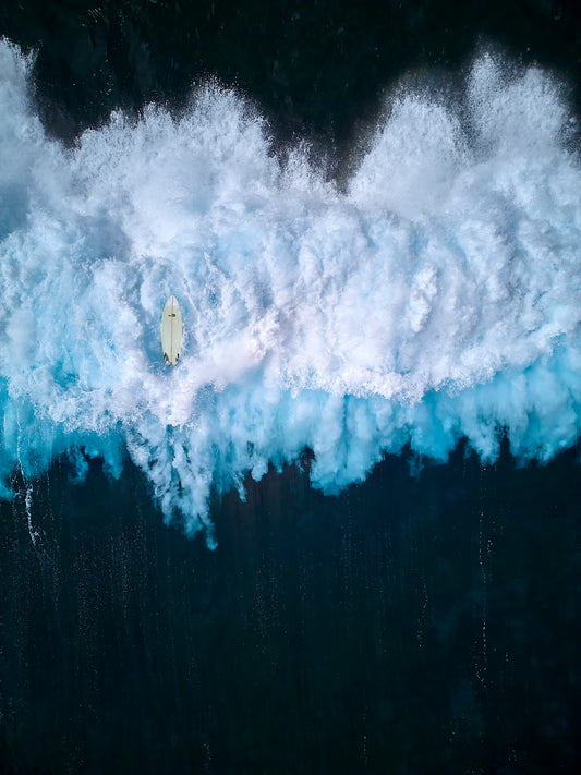 Aerial of foamy white wave crashing into dark blue surf, an upside down surfboard floats above the wave