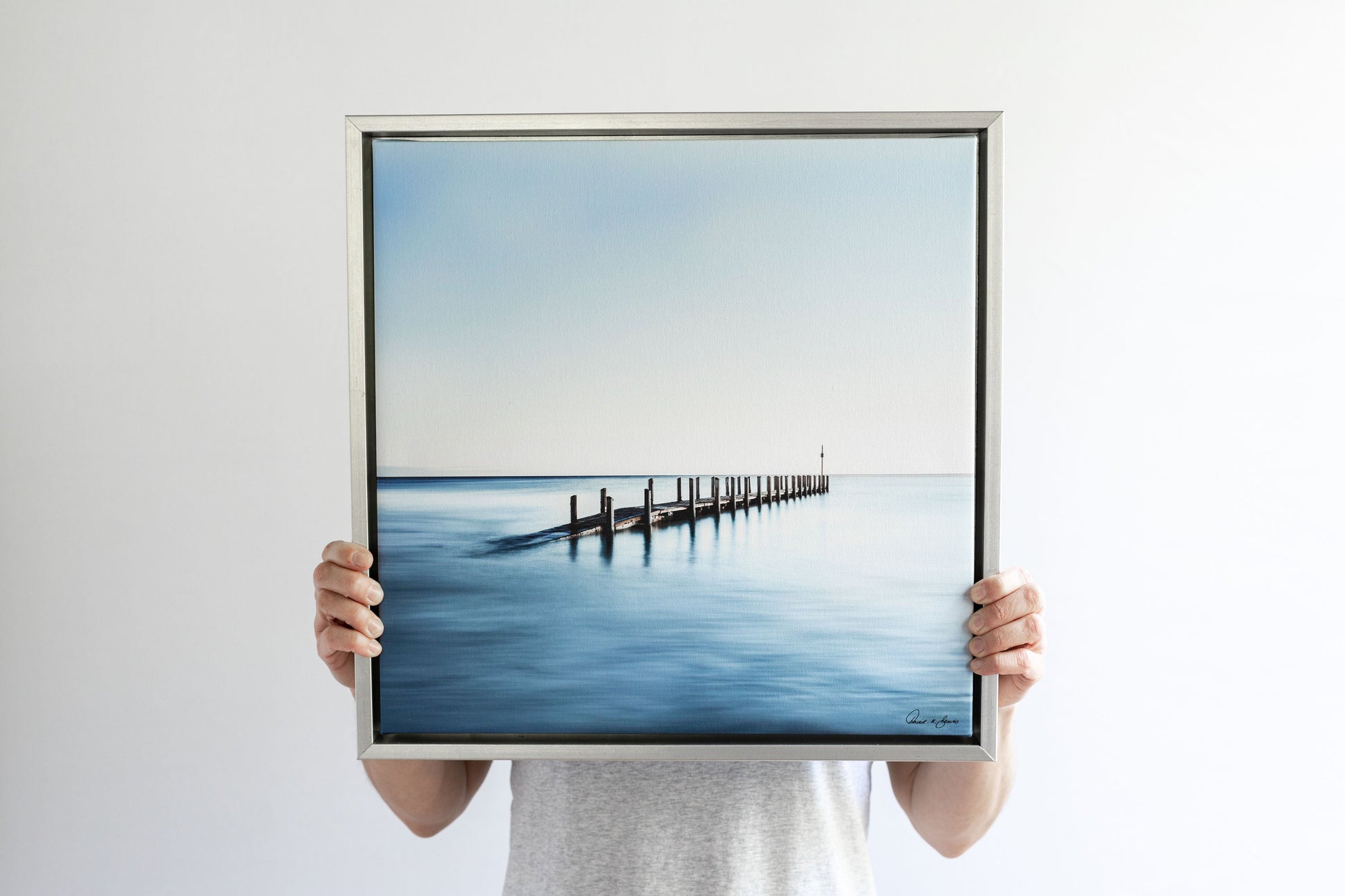 Person holding a framed fine art photograph. Grey and blue calm waters and wooden dock rising out of the ocean, David K Beavis Artist Choice Print High Tide